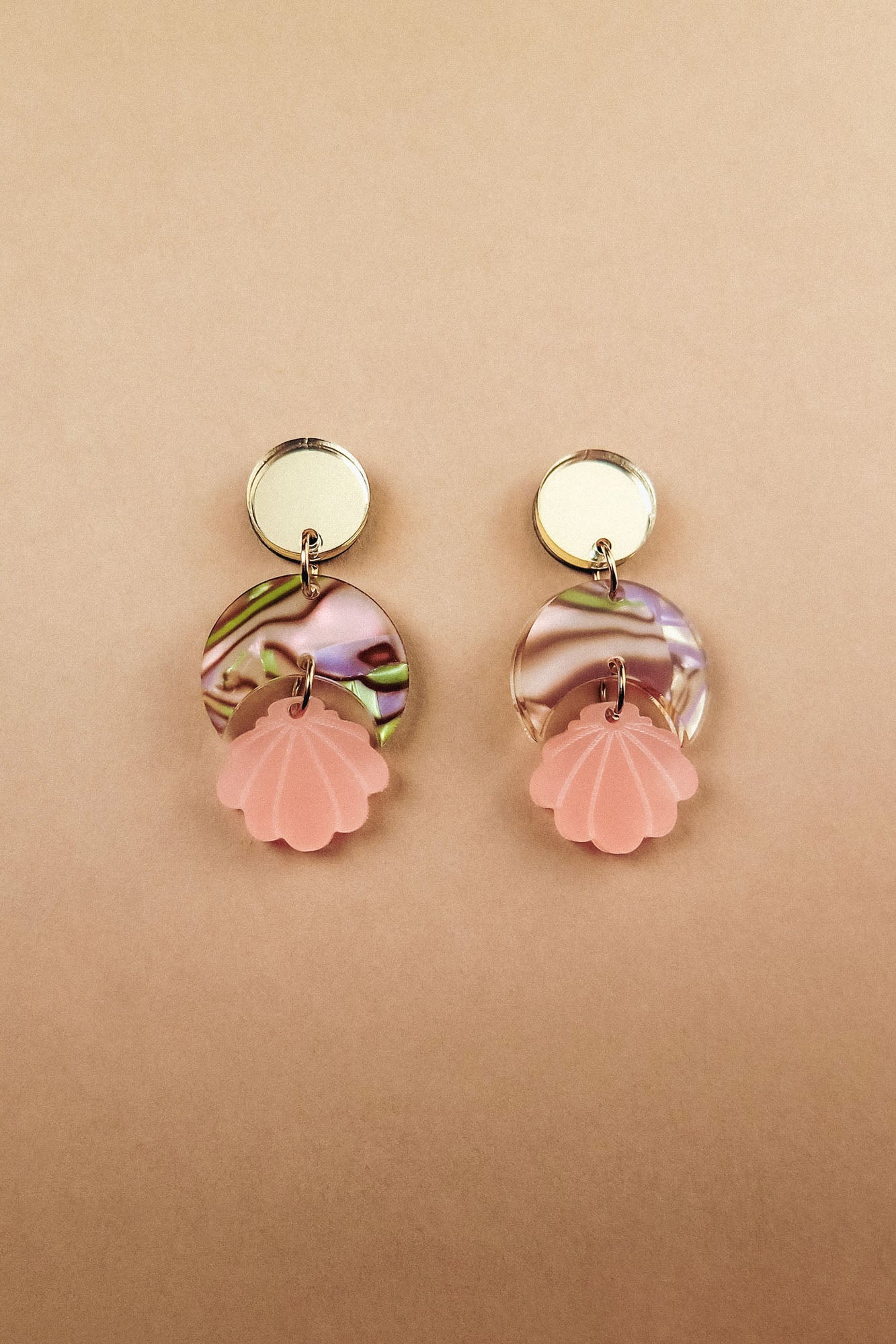 Handmade gold and marbled pink Seashell Earrings with circular tops, lightweight translucent marbled middles, and pink shell-shaped pendants, displayed on a light beige background.