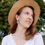 A woman with reddish-brown hair wearing a wide-brimmed straw hat, lightweight handmade seashell earrings, and a light V-neck top looks to the side, smiling softly. A blurred background of green trees is visible.