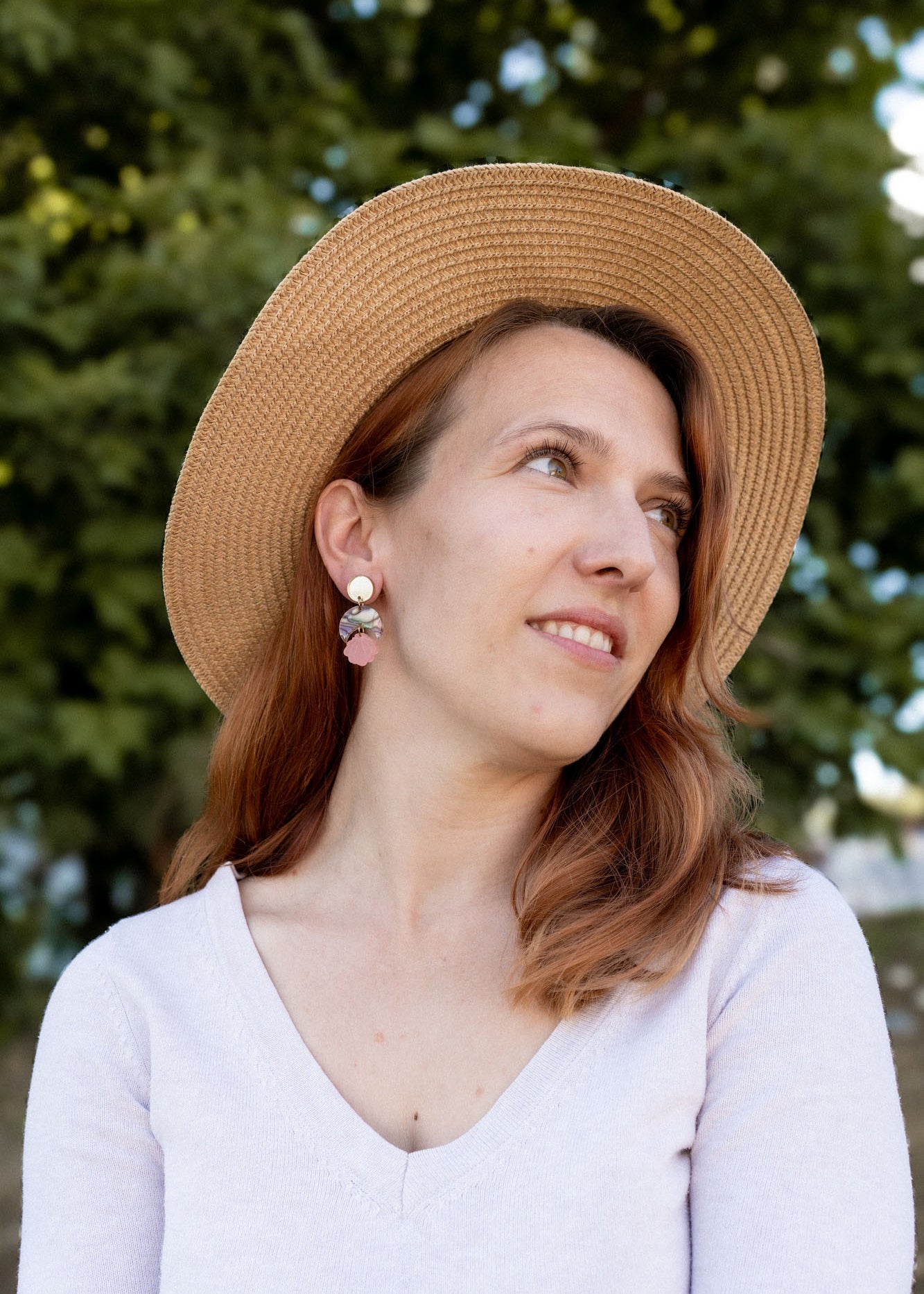 A woman with reddish-brown hair wearing a wide-brimmed straw hat, lightweight handmade seashell earrings, and a light V-neck top looks to the side, smiling softly. A blurred background of green trees is visible.