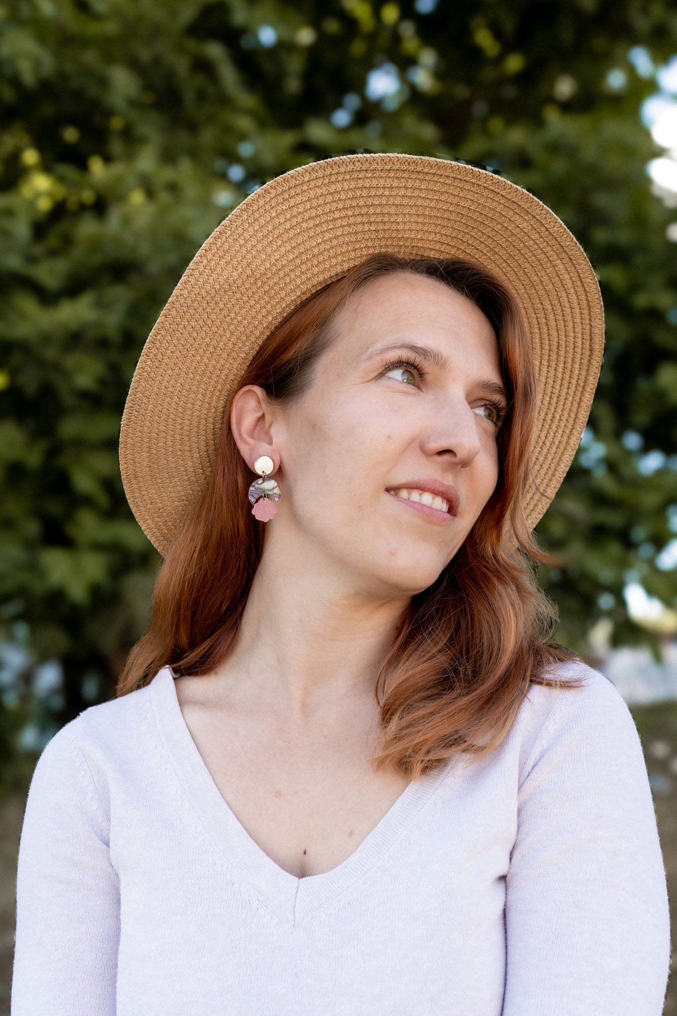 A woman with reddish-brown hair wearing a wide-brimmed straw hat, lightweight handmade seashell earrings, and a light V-neck top looks to the side, smiling softly. A blurred background of green trees is visible.