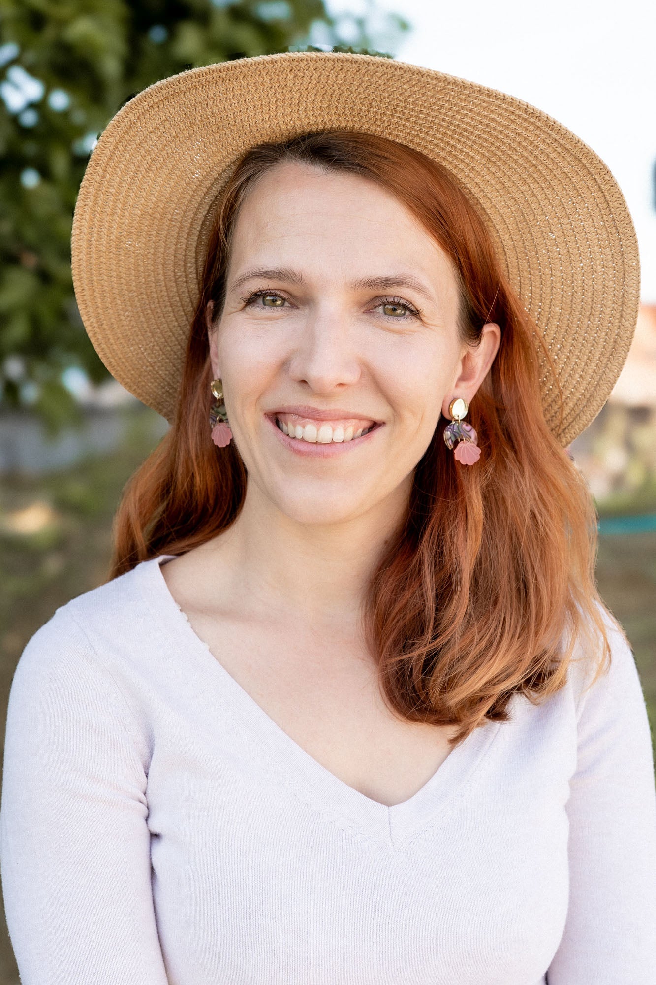 A woman with long auburn hair wearing a wide-brimmed straw hat, lightweight seashell earrings, and a light V-neck top smiles outdoors with greenery in the background.