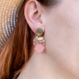 A close-up of a womans ear wearing a handmade gold stud earring with a bow and a dangling pink seashell-shaped charm. She has red hair, is wearing a straw hat, and the lightweight design adds delicate charm.