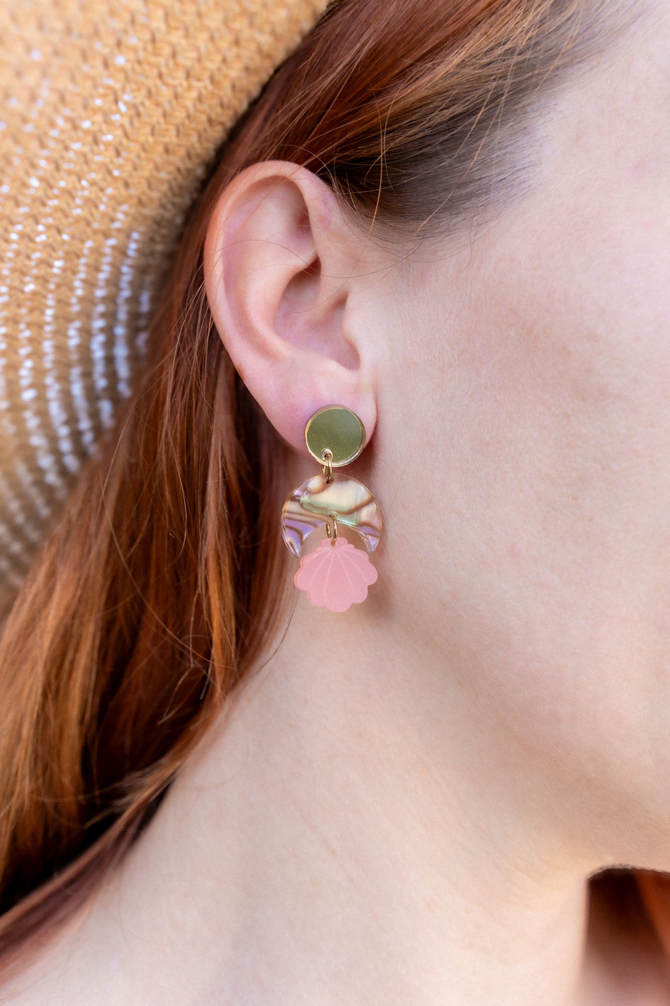 A close-up of a womans ear wearing a handmade gold stud earring with a bow and a dangling pink seashell-shaped charm. She has red hair, is wearing a straw hat, and the lightweight design adds delicate charm.