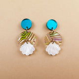 A pair of handmade seashell earrings featuring blue circular studs, marbled translucent discs with green and brown swirls, and white shell-shaped pendants, all lightweight and displayed on a beige background.
