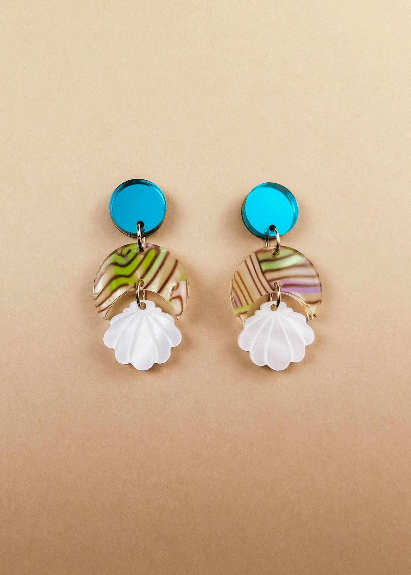A pair of handmade seashell earrings featuring blue circular studs, marbled translucent discs with green and brown swirls, and white shell-shaped pendants, all lightweight and displayed on a beige background.