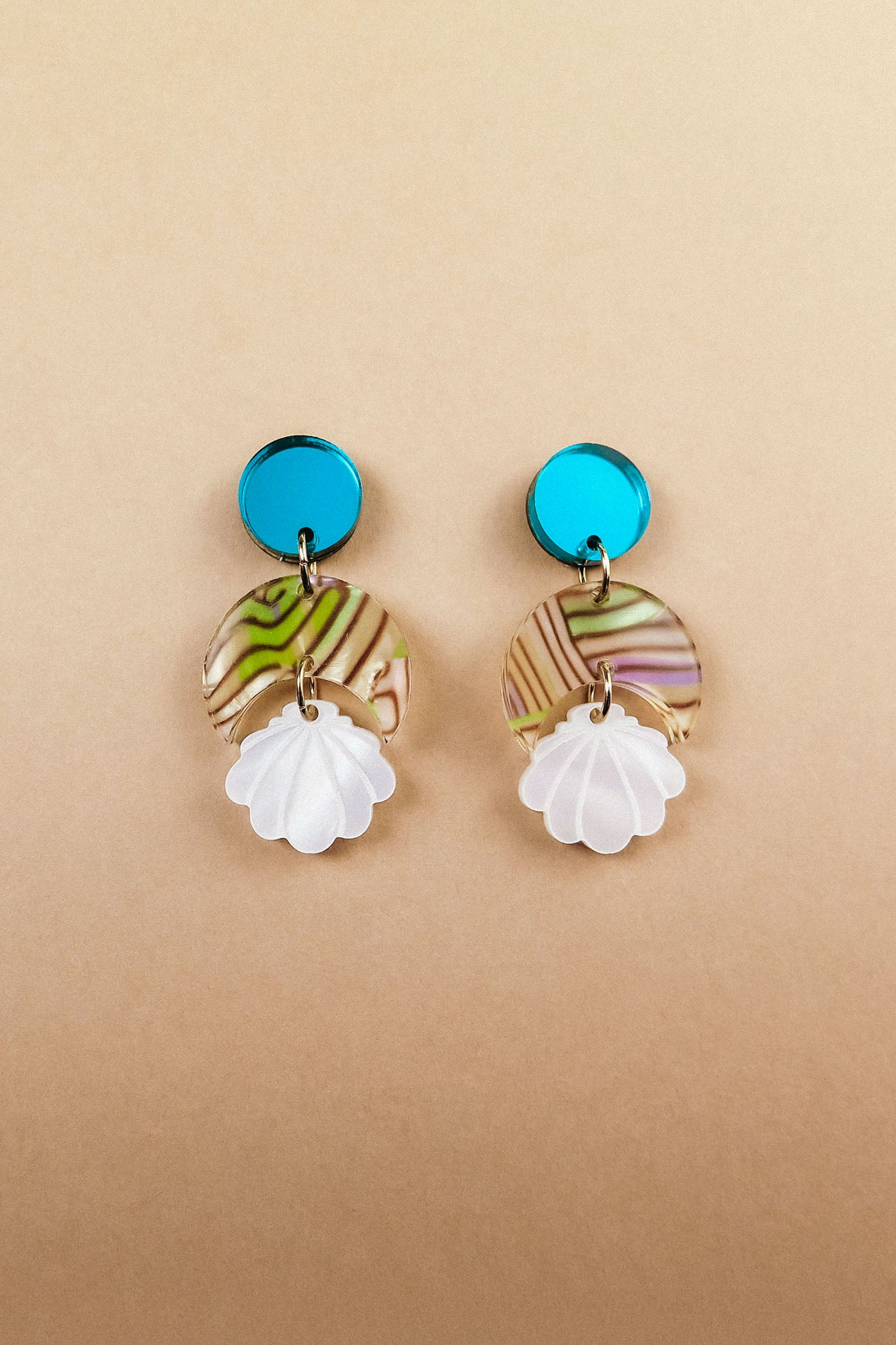 A pair of handmade seashell earrings featuring blue circular studs, marbled translucent discs with green and brown swirls, and white shell-shaped pendants, all lightweight and displayed on a beige background.