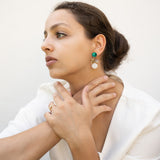 A woman with brown hair in a low bun wears a white blazer, gold rings, and lightweight seashell earrings with turquoise accents. She sits with her hands gently touching her neck, gazing thoughtfully to the side.