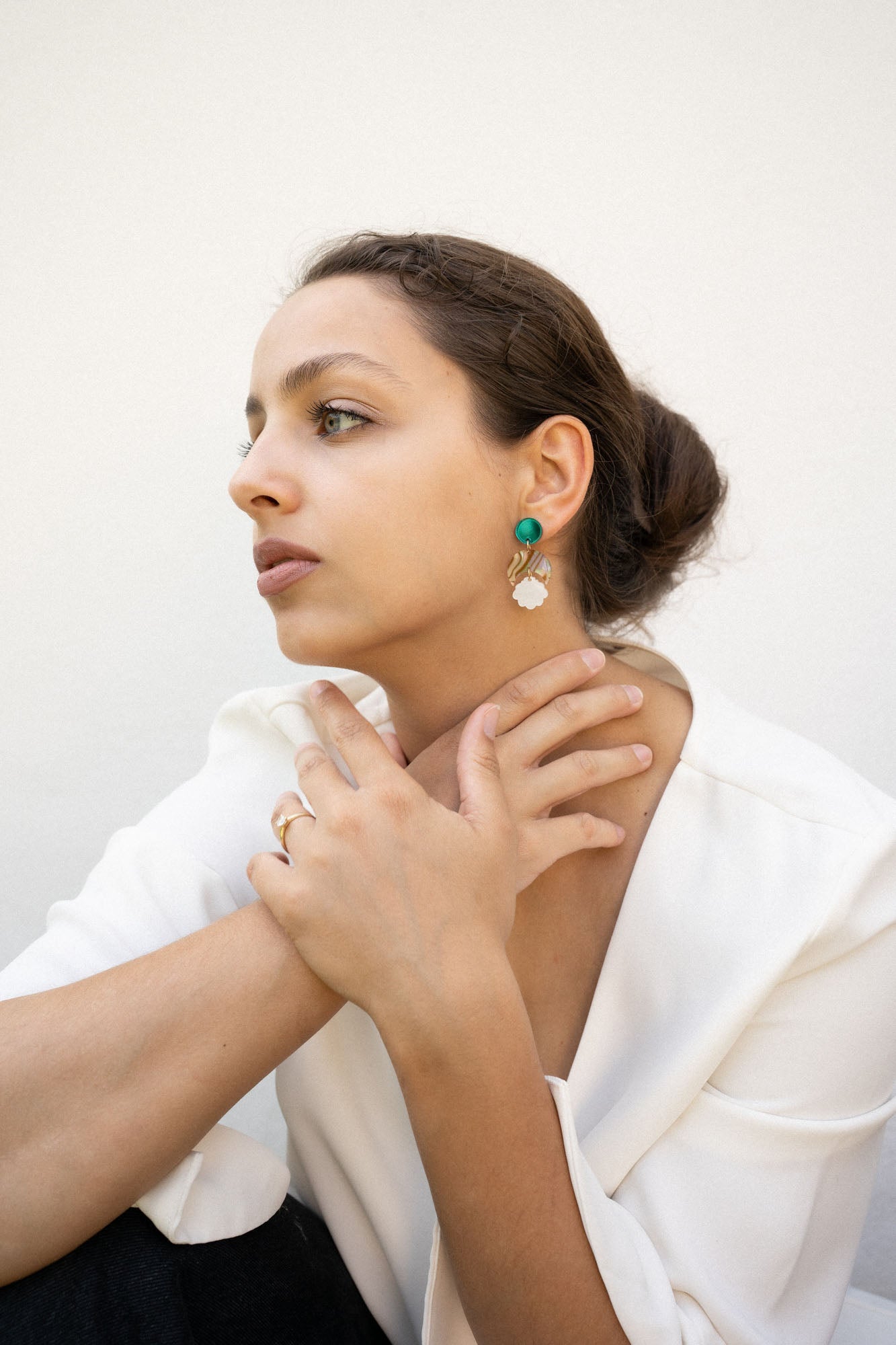 A woman with brown hair in a low bun wears a white blazer, gold rings, and lightweight seashell earrings with turquoise accents. She sits with her hands gently touching her neck, gazing thoughtfully to the side.
