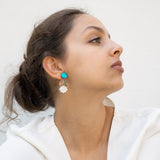 A woman with brown hair in a loose bun wears a white blouse and lightweight statement earrings with turquoise and gold accents. She gazes to the side against a plain, light background.