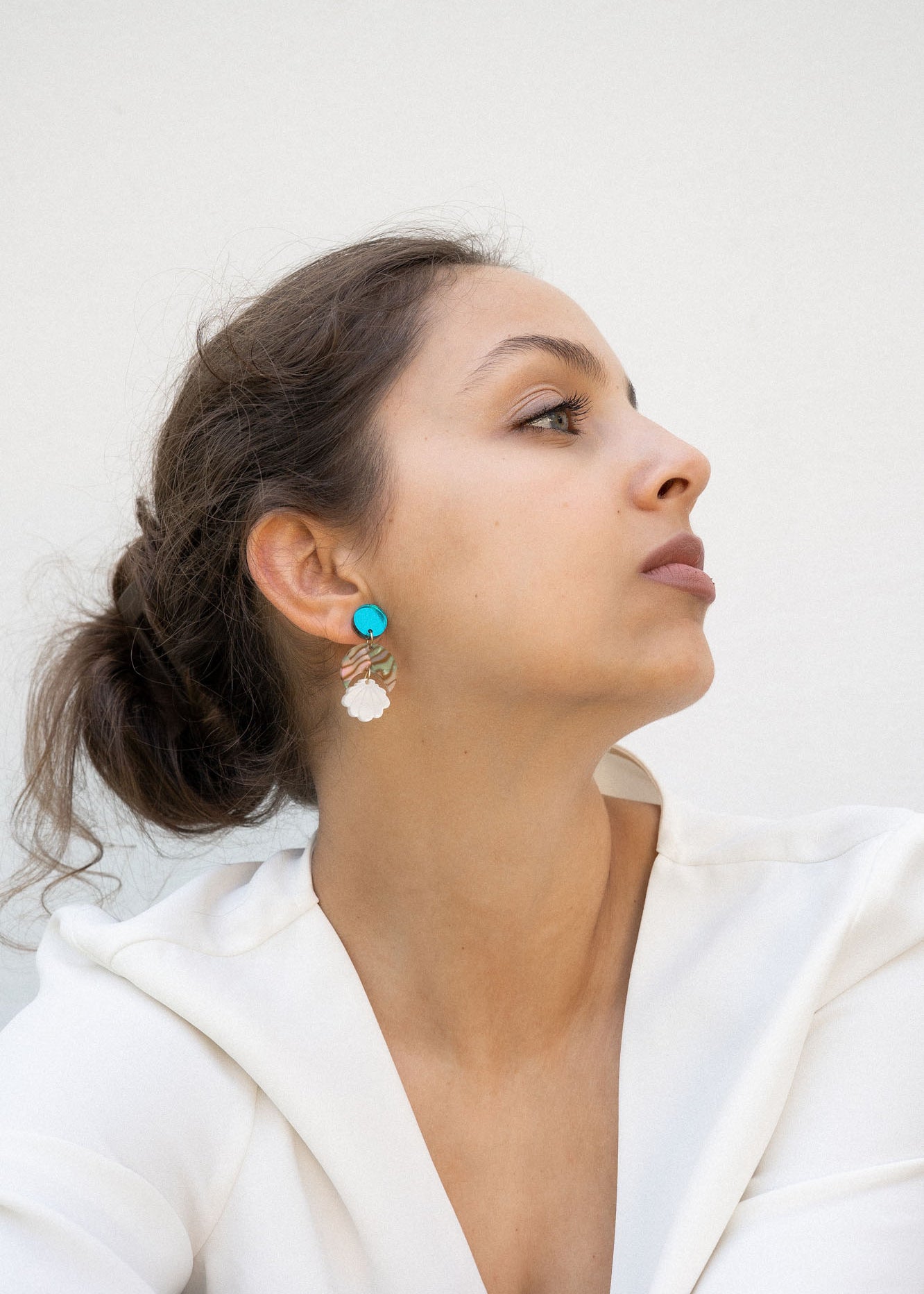 A woman with brown hair in a loose bun wears a white blouse and lightweight statement earrings with turquoise and gold accents. She gazes to the side against a plain, light background.