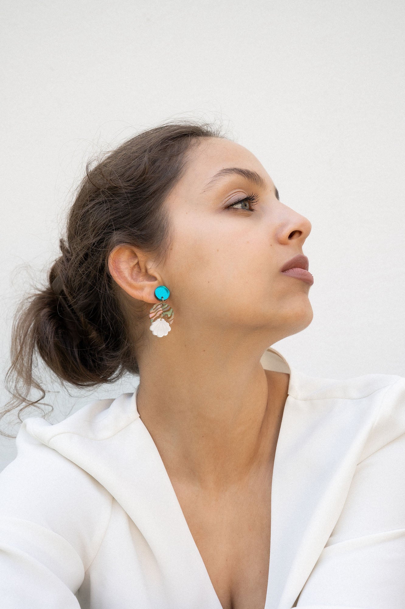 A woman with brown hair in a loose bun wears a white blouse and lightweight statement earrings with turquoise and gold accents. She gazes to the side against a plain, light background.