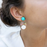 Close-up of a woman’s ear wearing a handmade, colorful earring with a turquoise stud, wavy striped section, and white floral-shaped dangle. Her hair is pulled back and she is wearing a white top. Lightweight for all-day comfort.