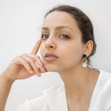 A woman with light skin and brown hair pulled back sits against a white background, wearing a white top and handmade seashell earrings. She rests her face on her hand, gazing thoughtfully at the camera.