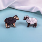 Two geometric wooden animal figures, one resembling a black sheep and the other a white sheep, are placed on a light blue surface with a soft white fabric backdrop, reminiscent of Polygon Sheep Stud Earrings adorned with sparkling rhinestones.