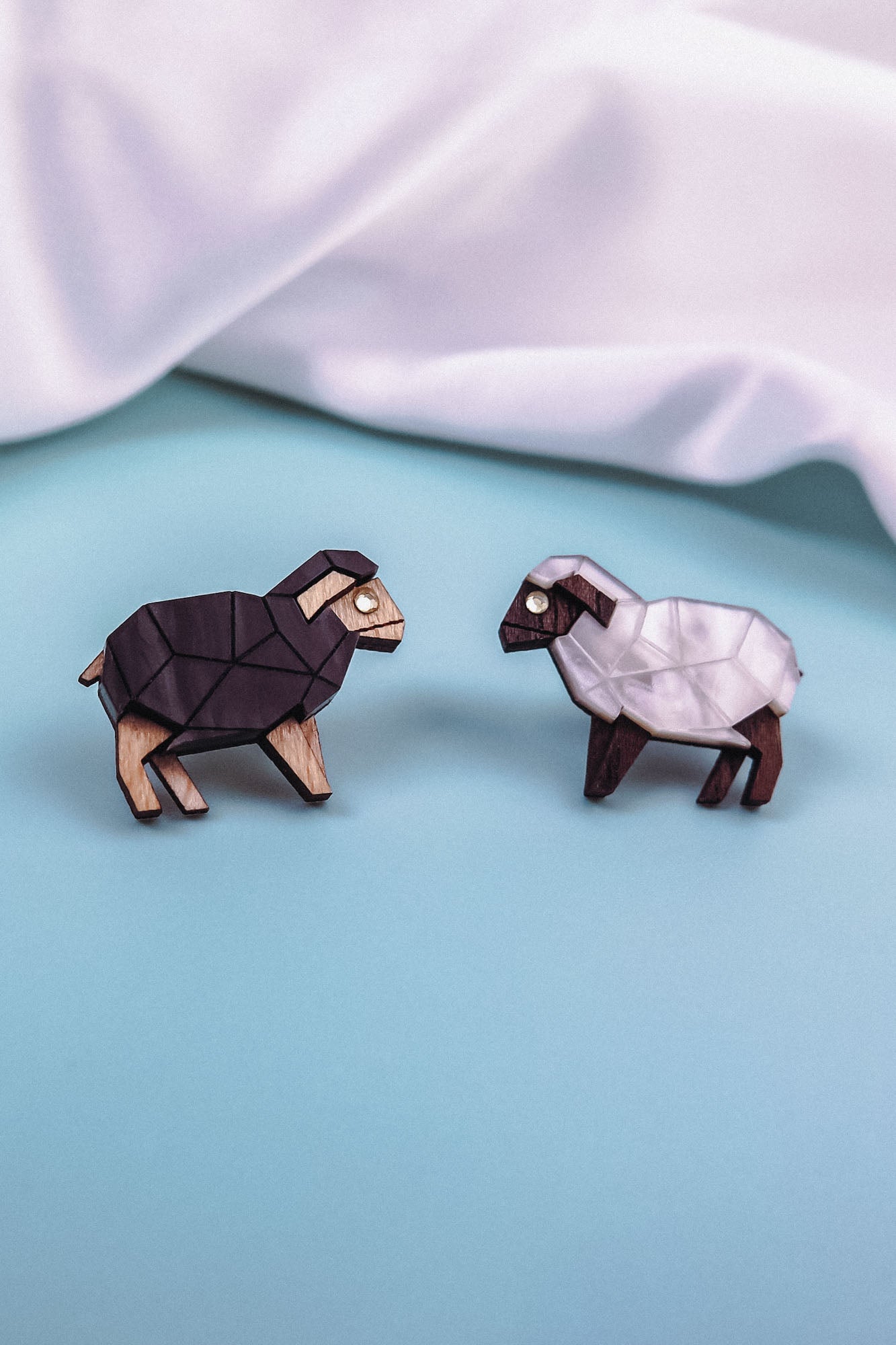 Two geometric wooden animal figures, one resembling a black sheep and the other a white sheep, are placed on a light blue surface with a soft white fabric backdrop, reminiscent of Polygon Sheep Stud Earrings adorned with sparkling rhinestones.