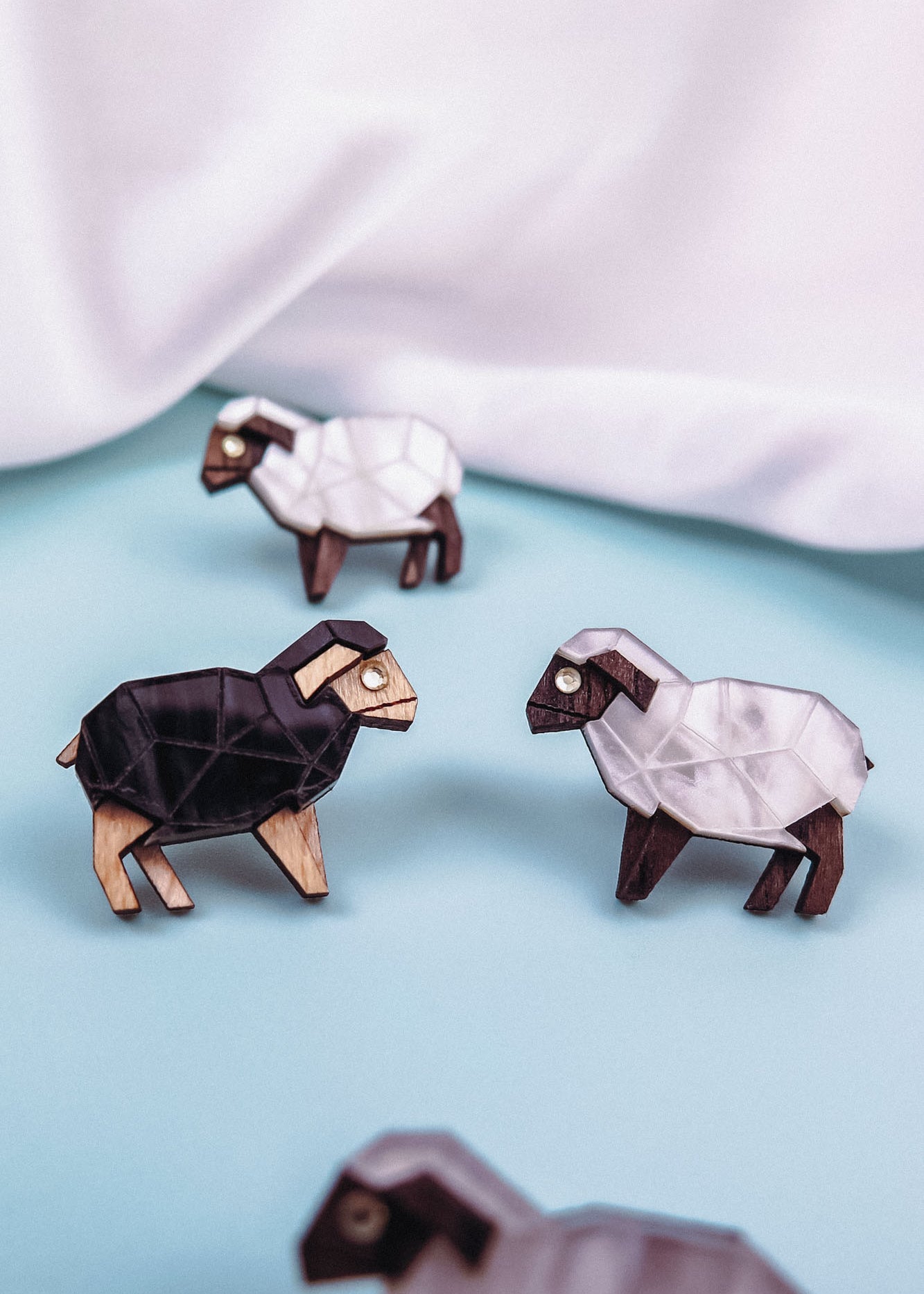 Four geometric wooden sheep figurines, three white and one black, are arranged on a light blue surface with a white fabric backdrop. Their polygonal shapes and minimalist design echo the style of Polygon Sheep Stud Earrings.