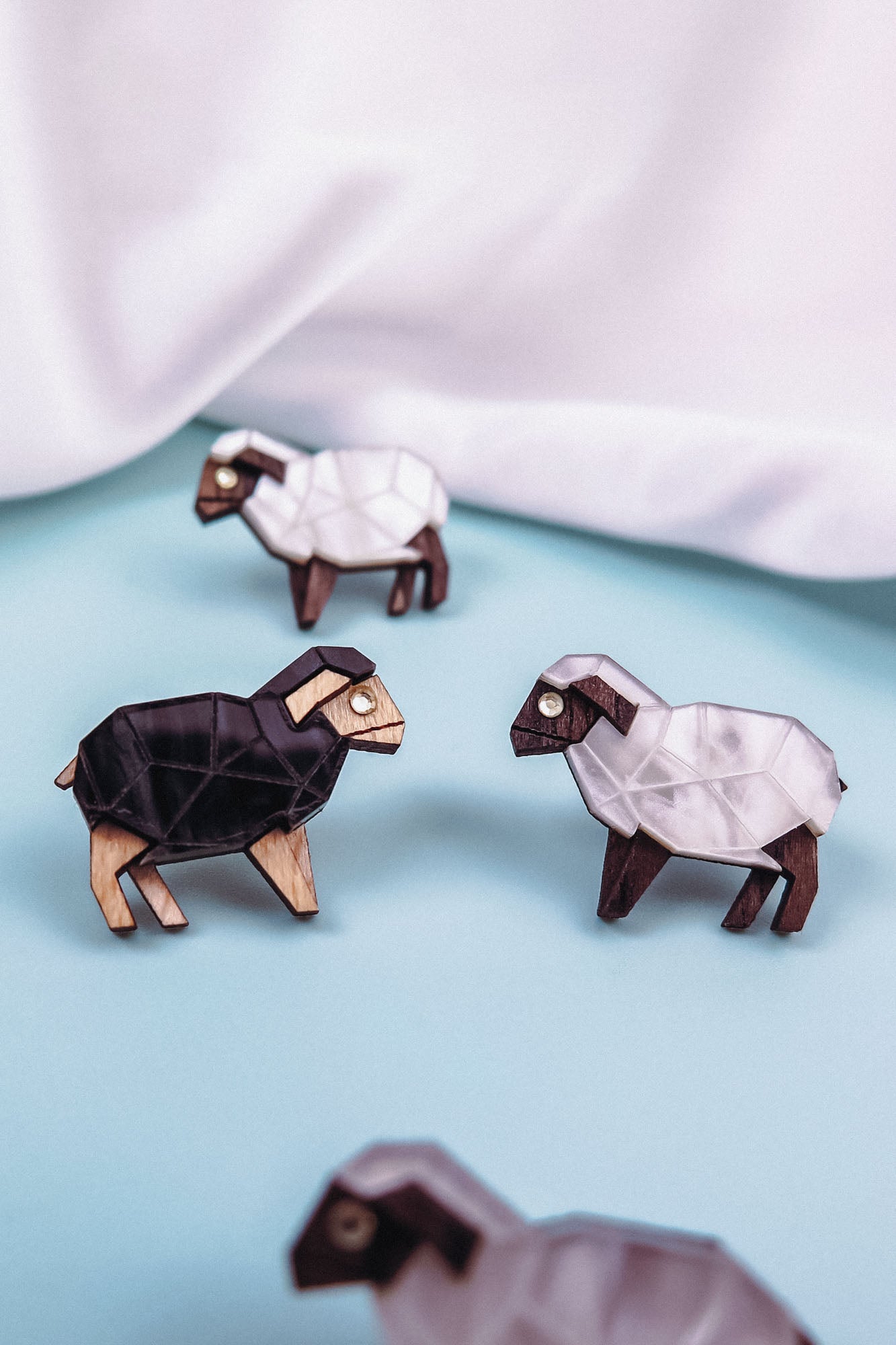 Four geometric wooden sheep figurines, three white and one black, are arranged on a light blue surface with a white fabric backdrop. Their polygonal shapes and minimalist design echo the style of Polygon Sheep Stud Earrings.