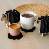 A white mug and a pink cup rest on felt coasters, accompanied by charming wooden sheep coaster holders on a light wooden table. Perfect as home decor accents, with a woven placemat in the background.