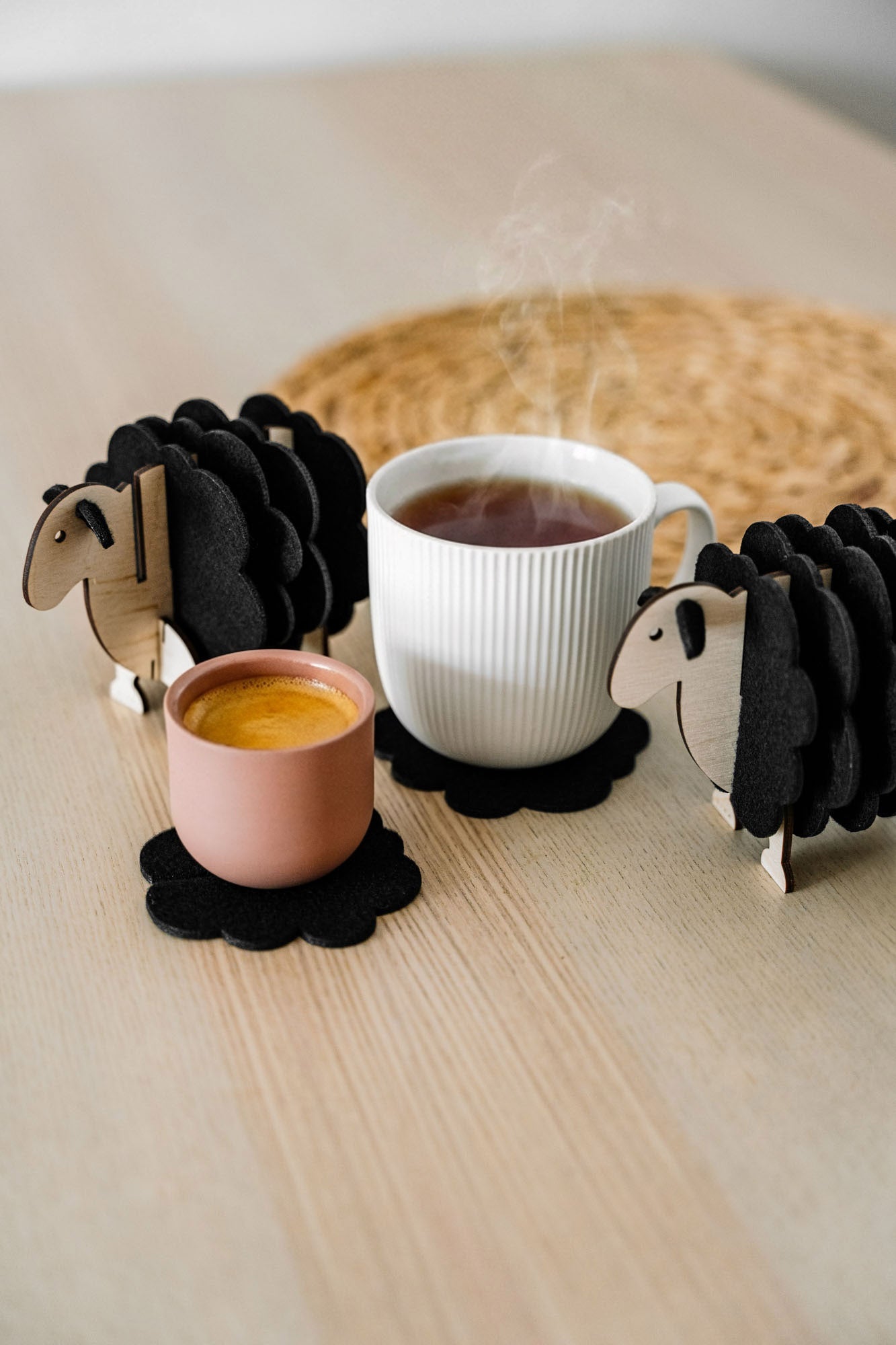 A white mug and a pink cup rest on felt coasters, accompanied by charming wooden sheep coaster holders on a light wooden table. Perfect as home decor accents, with a woven placemat in the background.