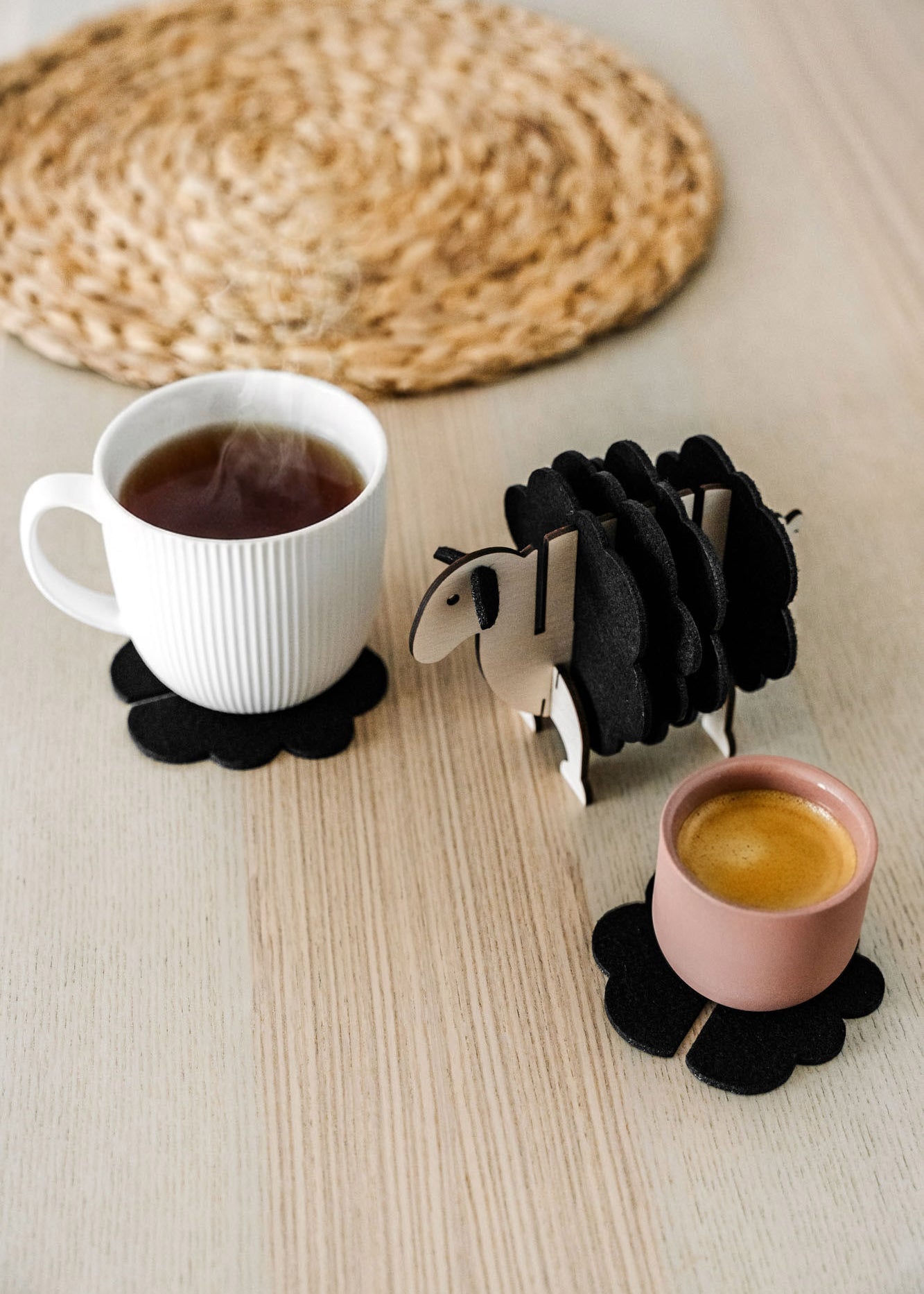 A white mug and a pink cup sit on black felt coasters next to a wooden sheep-shaped coaster holder on a light wooden table with a woven placemat in the background, perfect for charming home decor.