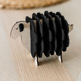 A charming wooden sheep-shaped coaster holder with black felt coasters stands on a light wooden table, bringing a touch of playful home decor alongside a woven placemat and white plate in the background.