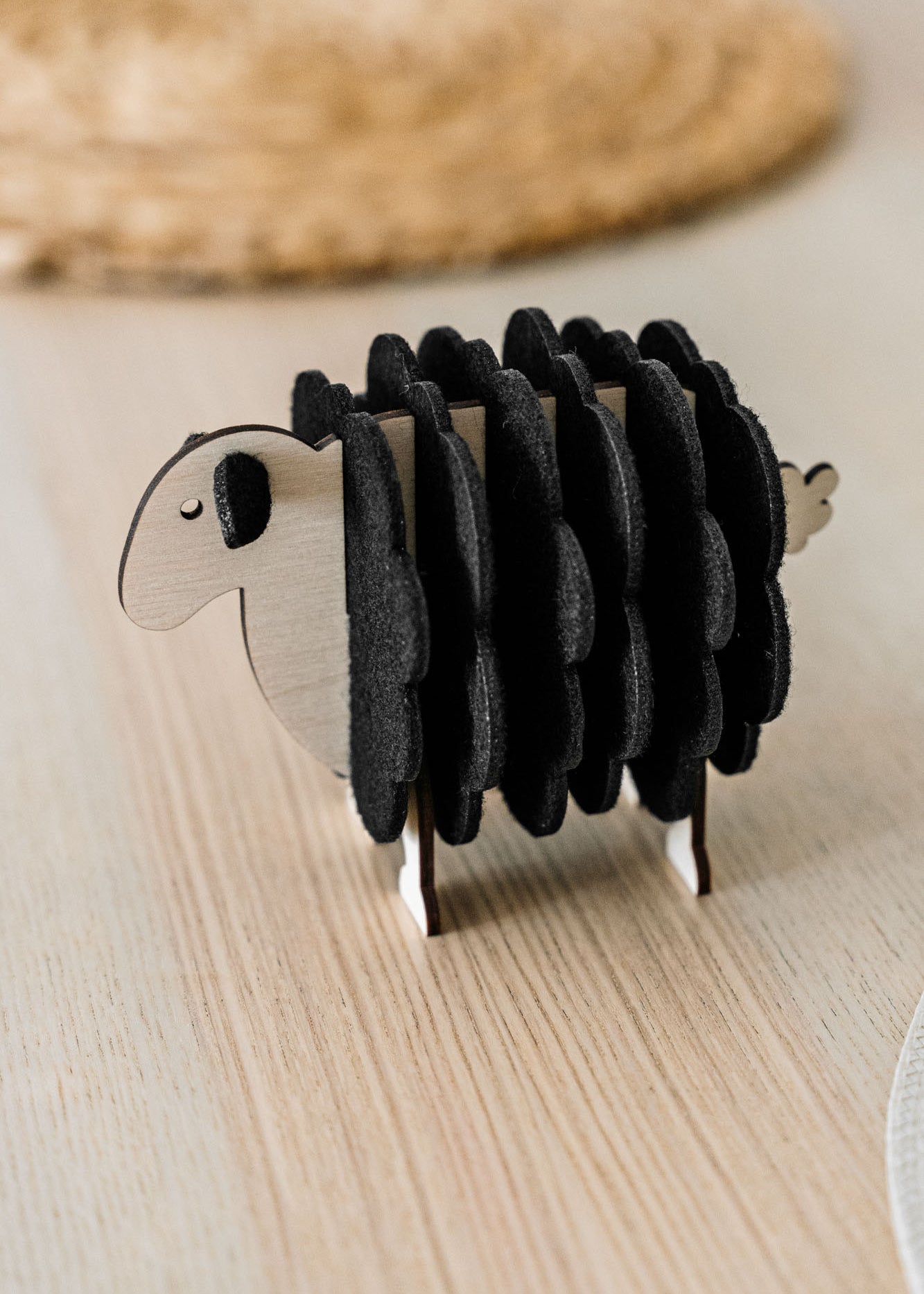 A charming wooden sheep-shaped coaster holder with black felt coasters stands on a light wooden table, bringing a touch of playful home decor alongside a woven placemat and white plate in the background.