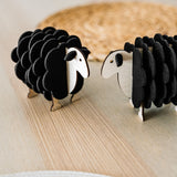 Two small, black wooden sheep figurines made of wood and felt stand facing each other on a light wooden surface, perfect for adding charm to your home decor, with a woven mat blurred in the background.