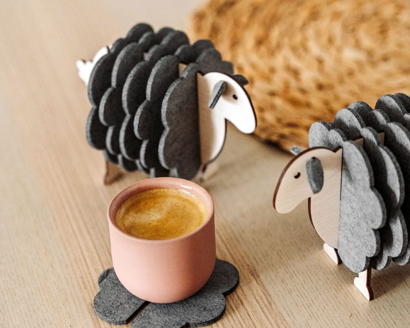 A pink cup rests on a gray, flower-shaped felt coaster beside two wooden sheep figurines—charming home decor—placed on a light wooden surface, with a woven mat partially visible in the background.