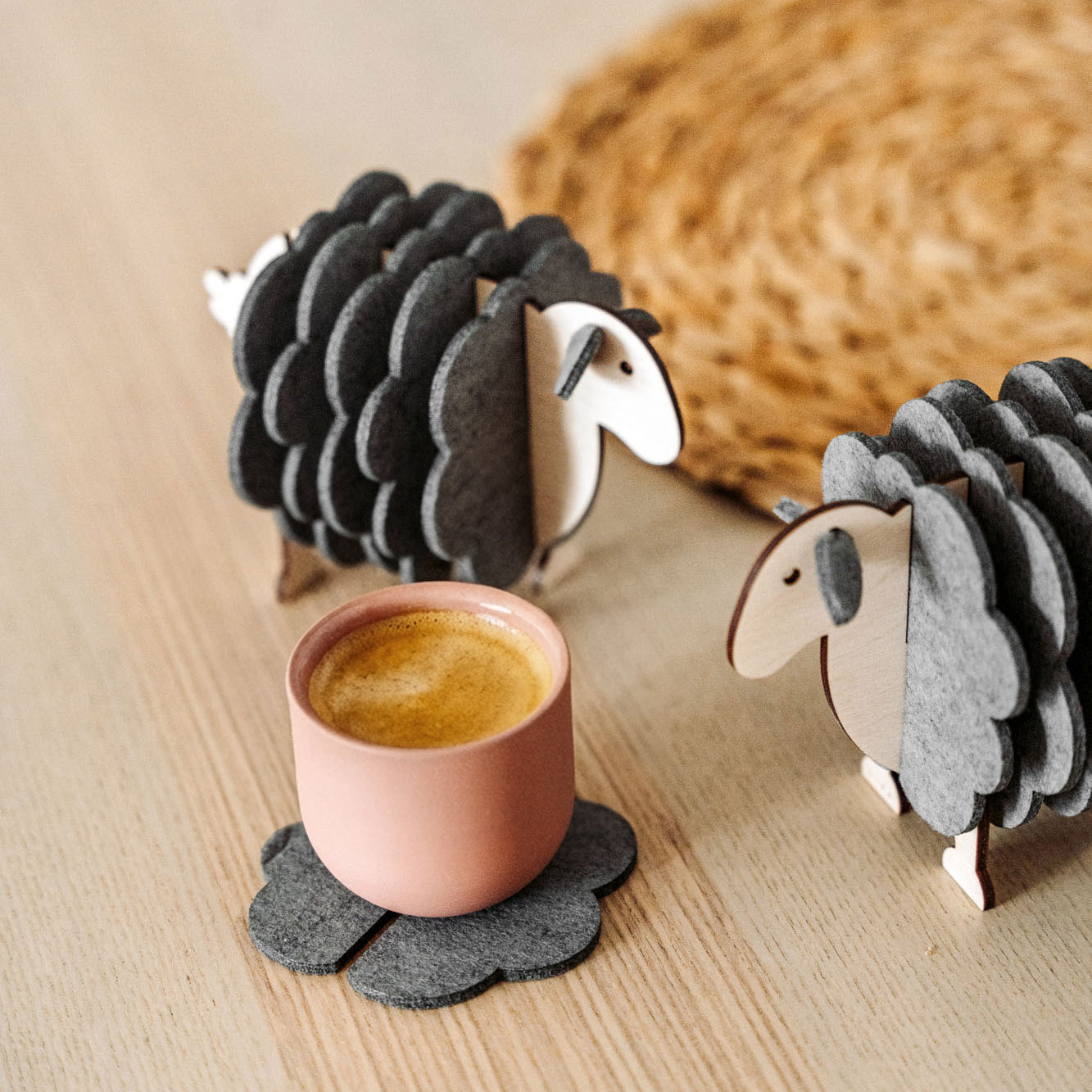 A pink cup rests on a gray, flower-shaped felt coaster beside two wooden sheep figurines—charming home decor—placed on a light wooden surface, with a woven mat partially visible in the background.