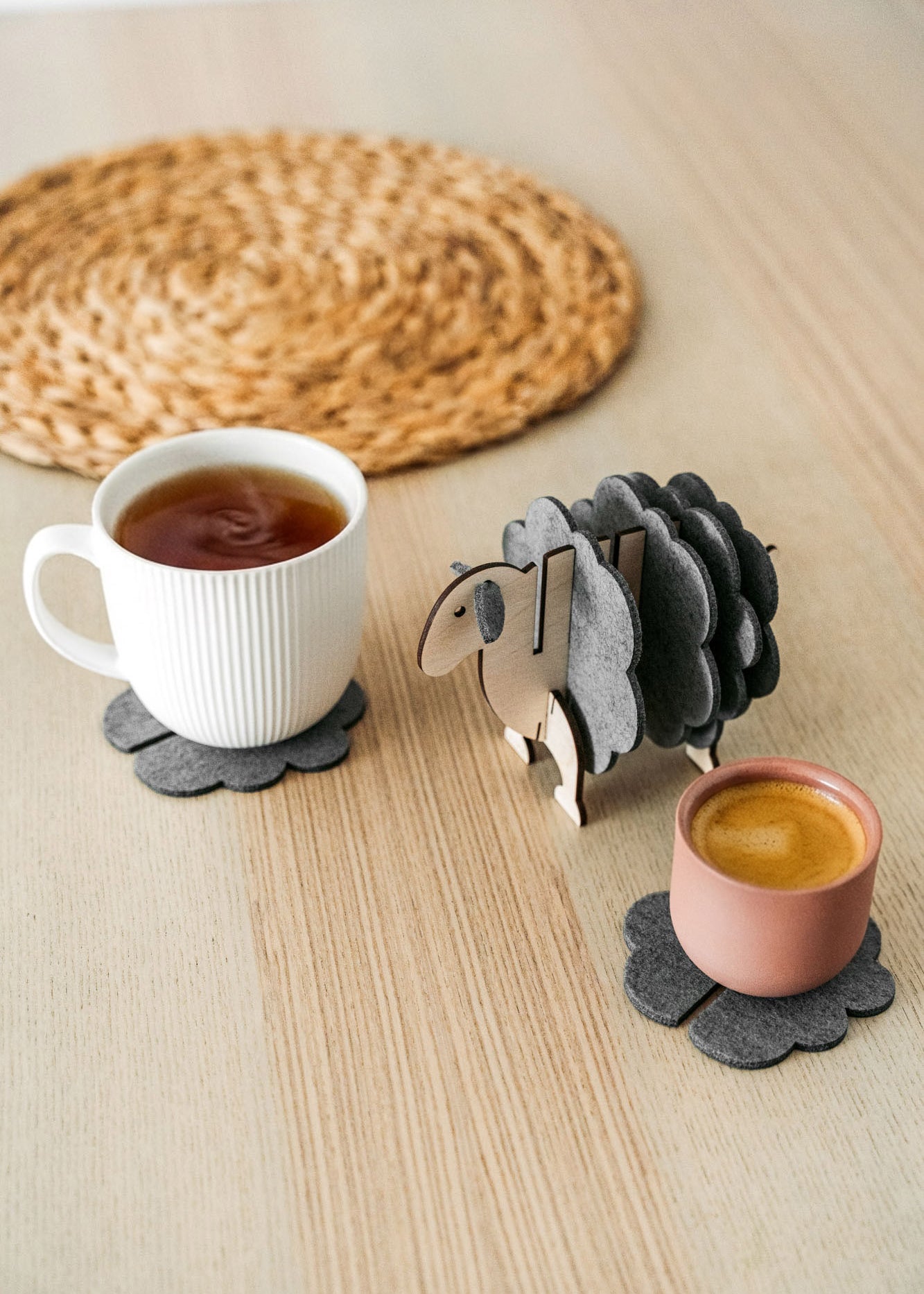 A white mug and a pink cup sit on dark felt coasters next to a decorative wooden sheep-shaped coaster holder, bringing charming home decor touches to a light wooden table topped with a round woven placemat.