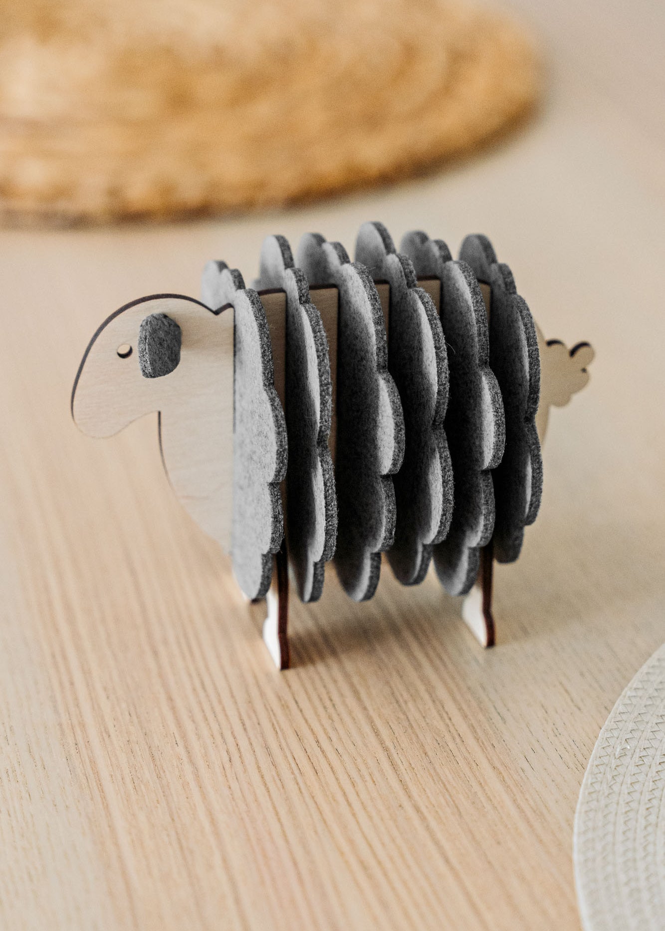 A wooden sheep-shaped coaster holder with round, gray felt coasters arranged to resemble the sheep’s wool stands on a light wooden table—perfect for charming home decor.
