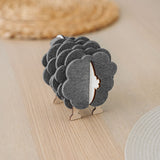 A wooden sheep-shaped coaster holder with dark felt coasters stands on a light table, adding a charming touch to your home decor. Woven placemats are visible in the background.