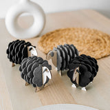 Three small, decorative wooden sheep figurines made of layered black and gray materials are arranged on a light wooden table with a woven placemat, perfect for adding charm to your home decor. A white vase sits in the background.