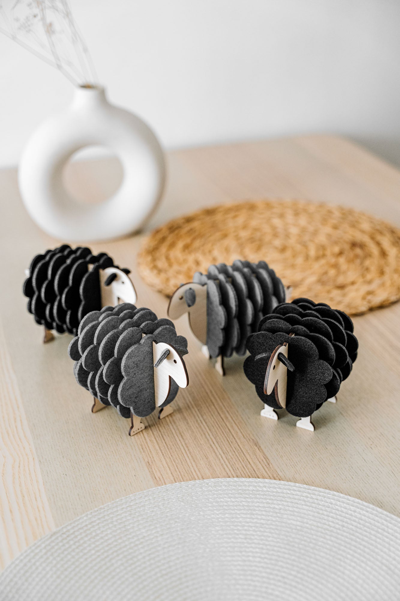 Three small, decorative wooden sheep figurines made of layered black and gray materials are arranged on a light wooden table with a woven placemat, perfect for adding charm to your home decor. A white vase sits in the background.