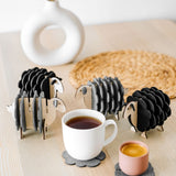 A white table holds a white vase, a woven placemat, and several sheep-shaped felt coasters. A white mug and a pink cup rest on two of the coasters, bringing playful charm to this cozy home decor setting.