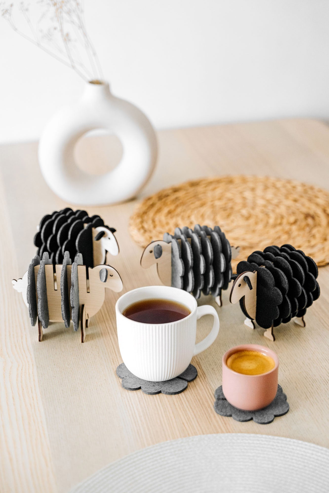 A white table holds a white vase, a woven placemat, and several sheep-shaped felt coasters. A white mug and a pink cup rest on two of the coasters, bringing playful charm to this cozy home decor setting.