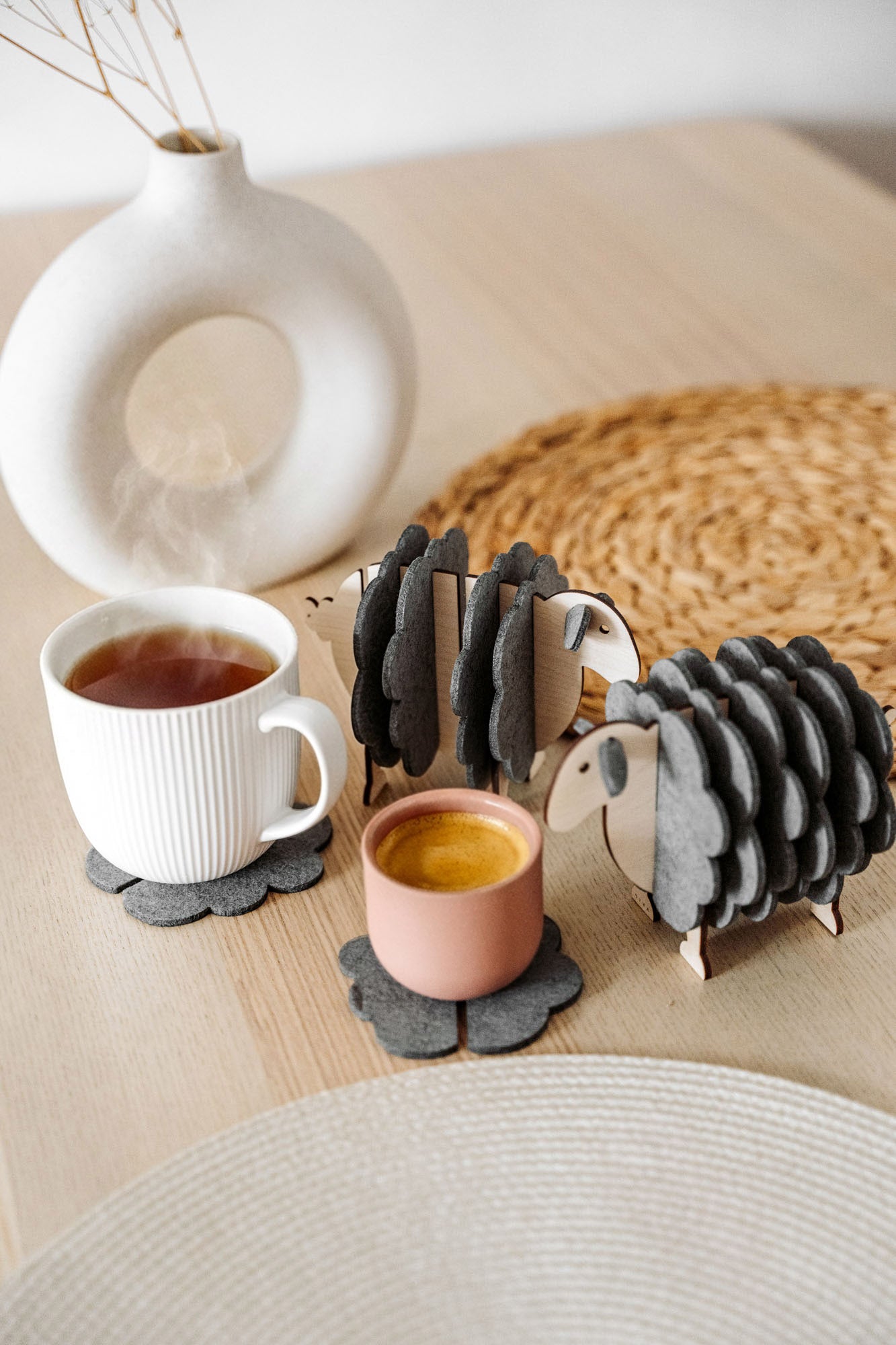 Two wooden sheep-shaped coaster holders with dark gray felt coasters add a touch of charming home decor to a light wooden table, paired with a white mug, pink cup, woven placemat, and modern white vase with dried stems.
