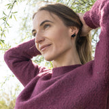 A woman in a purple sweater stands outdoors under tree branches, smiling gently and looking into the distance with her hands behind her head, sparkling rhinestones from her Polygon Sheep Stud Earrings catching the sunlight on a beautiful day.