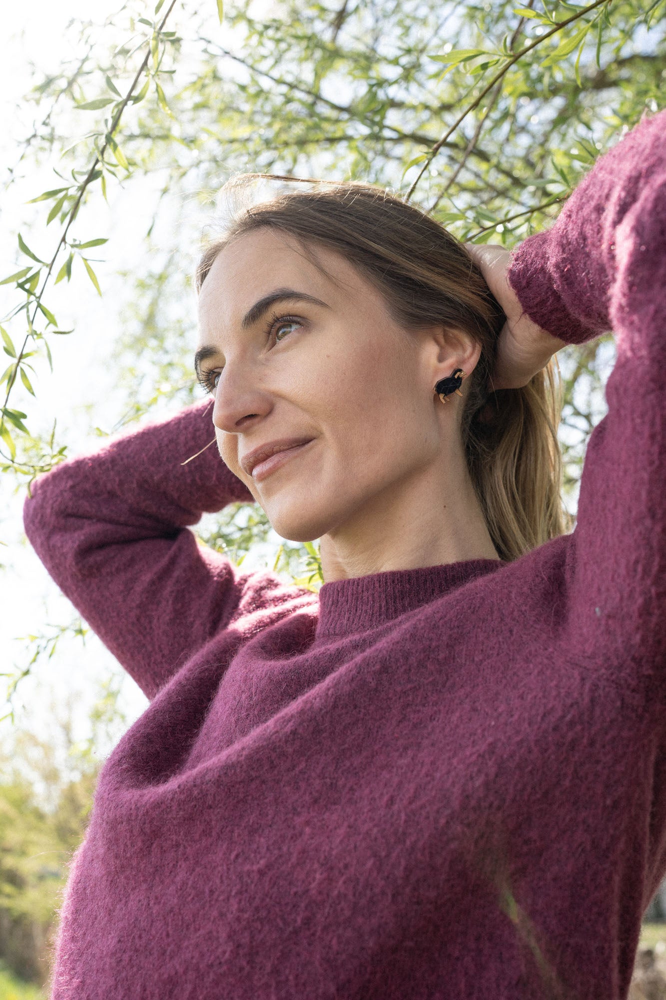 A woman in a purple sweater stands outdoors under tree branches, smiling gently and looking into the distance with her hands behind her head, sparkling rhinestones from her Polygon Sheep Stud Earrings catching the sunlight on a beautiful day.