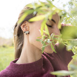 A woman in a maroon sweater stands outdoors with her eyes closed, partially obscured by green leaves, wearing handcrafted Polygon Sheep Stud Earrings adorned with sparkling rhinestones. The background is bright and blurred with greenery.
