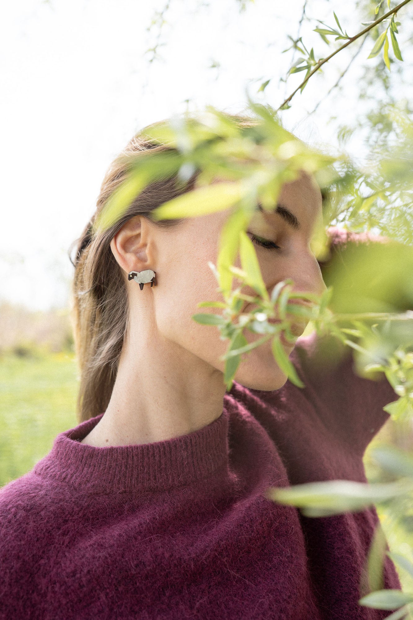 A woman in a maroon sweater stands outdoors with her eyes closed, partially obscured by green leaves, wearing handcrafted Polygon Sheep Stud Earrings adorned with sparkling rhinestones. The background is bright and blurred with greenery.