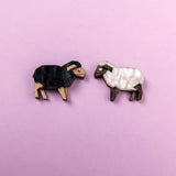 Two geometric, wooden sheep brooches—reminiscent of Polygon Sheep Stud Earrings—one black and one white—are placed on a light purple background, facing each other.