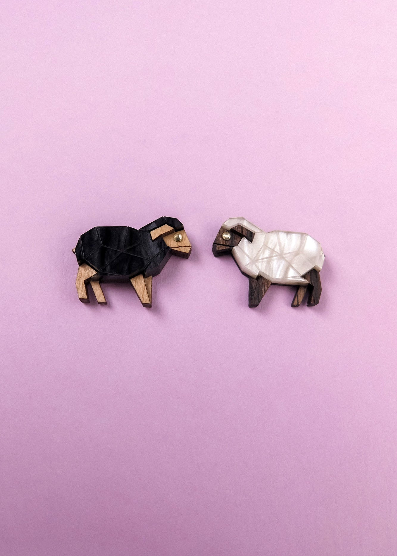 Two geometric, wooden sheep brooches—reminiscent of Polygon Sheep Stud Earrings—one black and one white—are placed on a light purple background, facing each other.