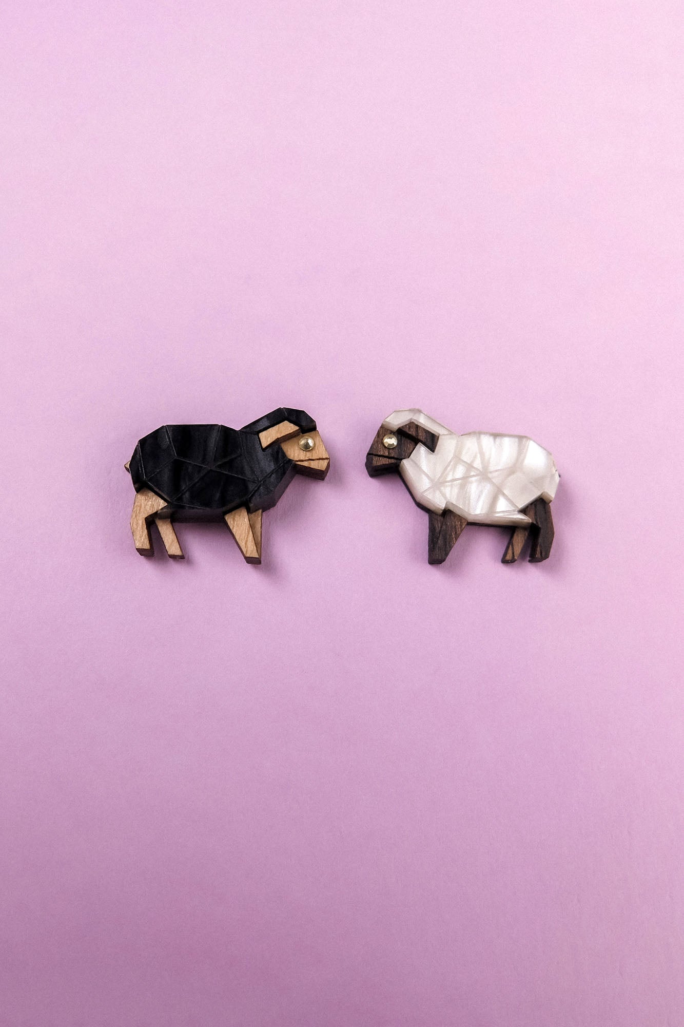 Two geometric, wooden sheep brooches—reminiscent of Polygon Sheep Stud Earrings—one black and one white—are placed on a light purple background, facing each other.