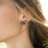Close-up of a person wearing a small, black and gold elephant-shaped handcrafted earring adorned with sparkling rhinestones. Their brown hair partially covers the ear, and they are wearing a burgundy top against a softly blurred background.