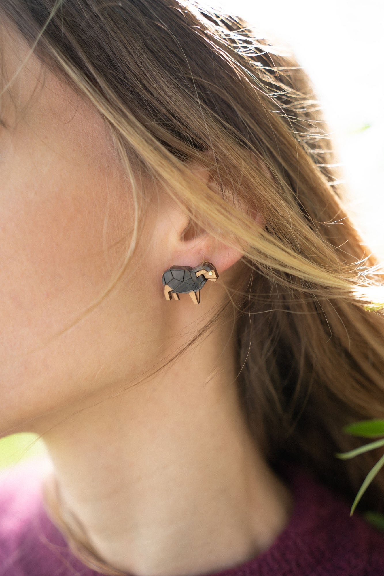 Close-up of a person wearing a small, black and gold elephant-shaped handcrafted earring adorned with sparkling rhinestones. Their brown hair partially covers the ear, and they are wearing a burgundy top against a softly blurred background.