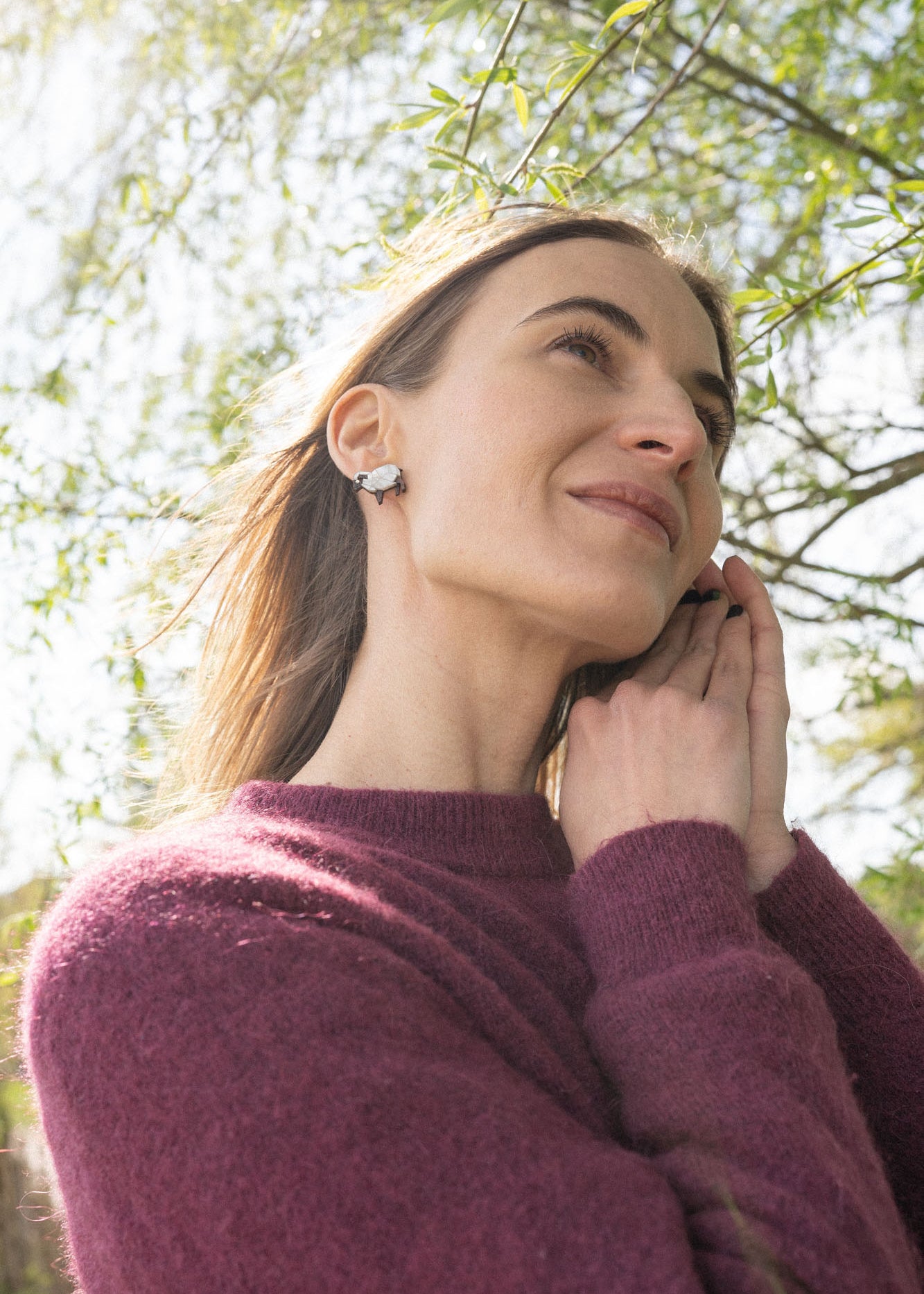 A woman with long brown hair wearing a purple sweater and handcrafted earrings stands outdoors, smiling softly as sunlight filters through the green leaves above her. Her hands gently touch her face, drawing attention to the sparkling rhinestones in her earrings.