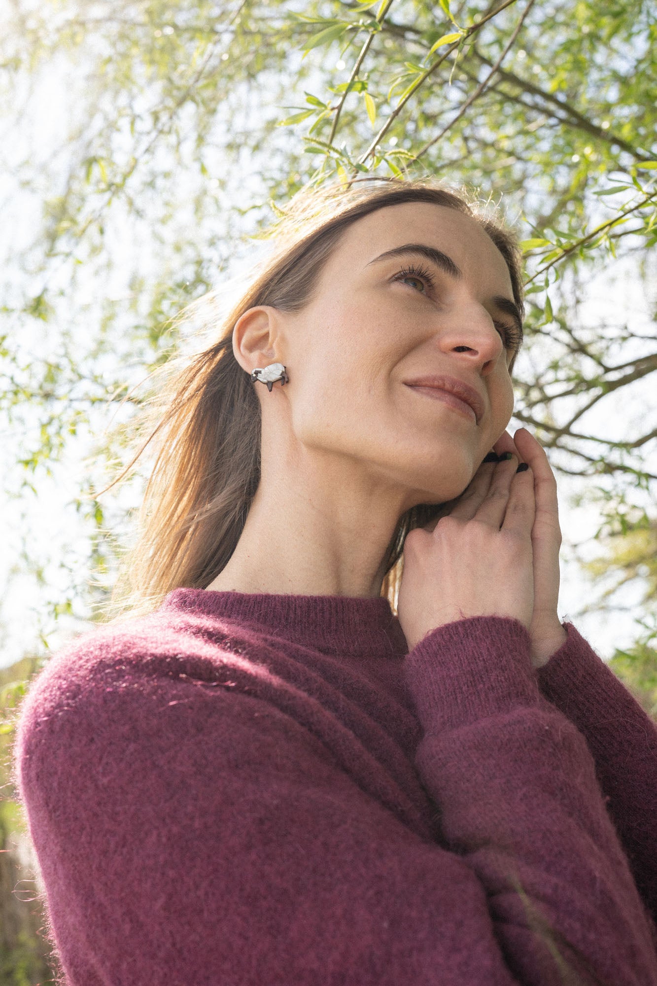 A woman with long brown hair wearing a purple sweater and handcrafted earrings stands outdoors, smiling softly as sunlight filters through the green leaves above her. Her hands gently touch her face, drawing attention to the sparkling rhinestones in her earrings.