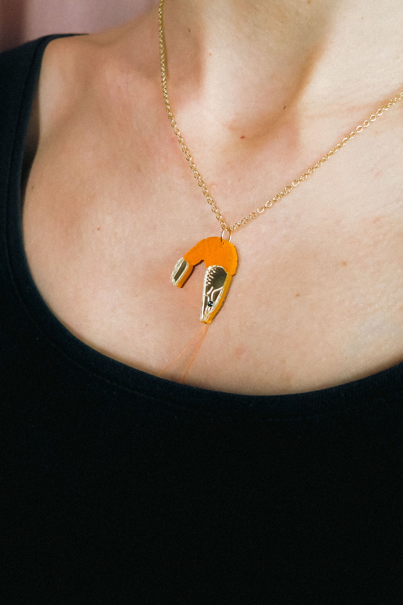 A whimsical Shrimp Pendant Necklace with a gold chain features an orange shrimp charm, playfully displayed above a black top on the wearer’s neck.