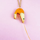 A whimsical Shrimp Pendant Necklace: this gold necklace features a playful orange shrimp pendant with a green gem eye and thin gold whiskers, beautifully displayed on a pastel pink background.